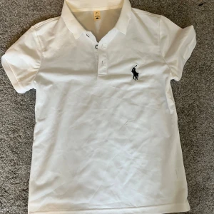 Vit pikétröja från Ralph Lauren - Vit pikétröja från Ralph Lauren med klassisk krage och korta ärmar. Tröjan har tre knappar framtill och den ikoniska svarta Polo-loggan broderad på bröstet. Perfekt för en clean och sportig look, tillverkad i mjuk bomull.