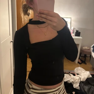 Svart långärmad polotopp med cut-out - Säljer en svart långärmad polotopp med cool cut-out detalj vid halsen och fyrkantig urringning. Toppen är tight och har en modern, edgy vibe som passar perfekt till jeans eller kjol. Materialet känns mjukt och stretchigt, förmodligen polyester eller bomull.