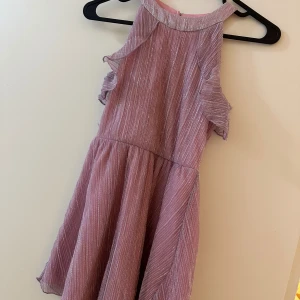 Glittrig rosa klänning från Lindex - Superfin rosa klänning från Lindex med glittriga ränder och volangdetaljer vid axlarna. Klänningen är ärmlös och har en lätt utställd kjol, perfekt för dig som gillar att sticka ut. Materialet är mjukt och skimrande, vilket ger en extra festlig känsla.