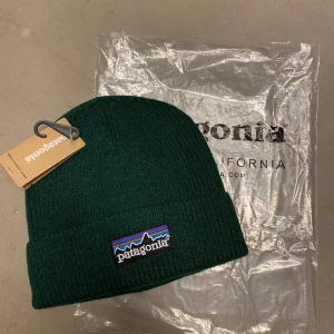 Mörkgrön ribbad mössa Patagonia - Snygg mörkgrön ribbstickad mössa från Patagonia med klassisk broderad logotyp framtill i färgglada toner. Perfekt passform och mjukt material som håller dig varm. En stilren accessoar för kalla dagar.
