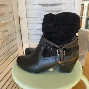 Svarta boots med snördetalj, storlek 38 - Svarta boots i konstskinn och mocka med dekorativt band och spänne runt ankeln. Snygg klack och rund tå, perfekt till höst och vinter. Bootsens ovandel är i mjuk mocka och nederdelen i slätt skinn. Stl 38 (EU).