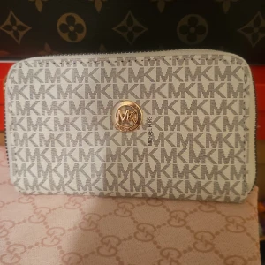 Vitplånbok med logomönster - Snygg plånbok  i vit och grå färg med MK-logomönster över hela ytan. Plånboken har guldfärgade detaljer, dragkedja och ett stort MK-emblem framtill. Insidan har flera fack för kort och en dragkedja för mynt. Perfekt för dig som gillar stilrena accessoarer.