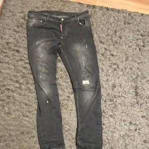 Gråa Dsquared2 jeans  - Säljer ett par svarta jeans från Dsquared2 med coola slitningar och färgstänk i rött och vitt. Jeansen har smal passform, klassisk femficksdesign och tydlig Dsquared2-patch bak. Materialet är jeans och de har en modern, streetig vibe.