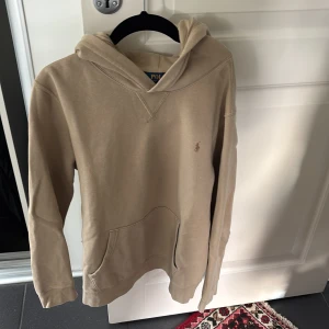 Beige hoodie från Polo Ralph Lauren - Snygg beige hoodie från Polo Ralph Lauren i storlek s. Tröjan har huva, känguruficka och det klassiska lilla Polo-logot på bröstet. Tillverkad i mjuk bomull, perfekt för chill dagar. Långärmad och med avslappnad passform.