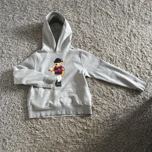 Grå hoodie Polo Ralph Lauren L - Säljer en grå hoodie från Polo Ralph Lauren i storlek 14-16år. Tröjan har ett stort tryck med den klassiska Polo-björnen på framsidan, huva och långa ärmar. Mjuk insida och ribbade muddar. Perfekt för dig som gillar sportig och chill stil.