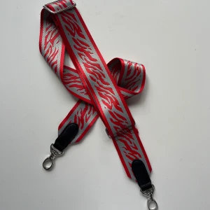 Justerbar axelrem Beck Söndergaard - Justerbart axelband till väska från Beck Söndergaard. Röd och grå zebra/tiger-mönster, tror att modellen heter zestroke bag strap. Använt fåtal gånger, i jättefint skick. Längd justerbar i intervallet 76-122 cm.
