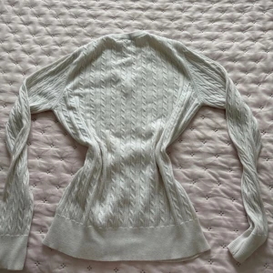 Vit kabelstickad tröja Tommy Hilfiger - Kabelstickad vit tröja från Tommy Hilfiger med v-ringning och diskret logga på bröstet. Tröjan har lång ärm, ribbade muddar och är gjord i mjuk bomull. Perfekt för lager-på-lager och snygg till jeans eller kjol.