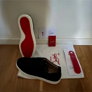 Svarta sneakers från Christian Louboutin - Säljer ett par svarta sneakers från Christian Louboutin med klassisk röd sula och vit kant. Skorna har en stilren design i mocka och är perfekta för dig som vill sticka ut med exklusiva detaljer. Kommer med originalpåse och tillbehör.