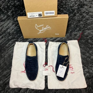 Mörkblå Christian Louboutin sneakers - Säljer ett par mörkblå sneakers från Christian Louboutin i mocka med ikoniska röda sulor. Skorna har snörning och coola svarta nitar på tån för en edgy vibe. Insidan är i beige skinn och yttersulan är vit. Perfekta för dig som vill sticka ut.