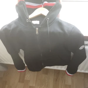 Svart hoodie från Moncler med dragkedja - Svart hoodie från Moncler med hel dragkedja, huva med snörning och Moncler-logga på ärmen. Muddarna har snygga ränder i rött, vitt och blått. Perfekt för dig som gillar stilrena och sportiga plagg.