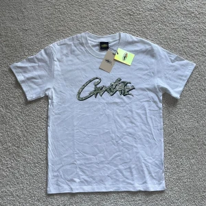 Vit Corteiz t-shirt med camo-logga - Vit t-shirt från Corteiz med stor camo-inspirerad logga på bröstet. Klassisk passform med rund hals och korta ärmar. Tillverkad i mjuk bomull och har autentiska tags kvar. Perfekt för dig som gillar streetwear och vill sticka ut.