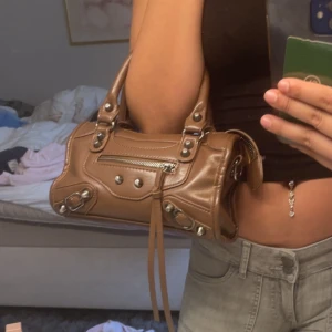 Brun handväska med silverdetaljer - Snygg brun handväska i skinn med coola silverdetaljer och dragkedja framtill. Väskan har två handtag och dekorativa nitar samt långa remmar som hänger ner. Perfekt storlek för det viktigaste och har en trendig, lite edgy look.