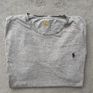 Grå t-shirt från Polo Ralph Lauren - Snygg grå t-shirt från Polo Ralph Lauren med klassisk passform och rund hals. T-shirten har det ikoniska broderade logotypen på bröstet och är tillverkad i mjuk bomull. Perfekt basic-plagg med stilren look.