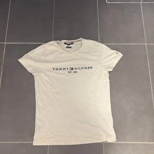 Vit Tommy Hilfiger t-shirt med tryck - Vit t-shirt från Tommy Hilfiger med klassiskt tryck på bröstet och logga på ärmen. Rund hals och korta ärmar. Tillverkad i mjuk bomull som känns skön mot huden. Snygg och enkel design som funkar till jeans eller shorts.