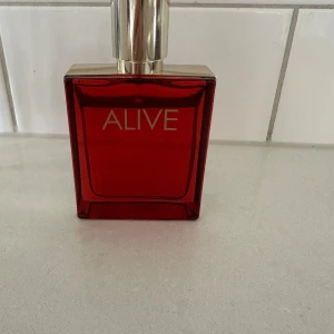 Hugo Boss Alive parfym - Stilren röd flaska med guldlock och tydlig 'ALIVE'-logga. Perfekt för dig som vill ha en modern och trendig parfym från Hugo Boss. Passar dig som gillar att sticka ut och vill ha något som känns fräscht och energiskt.