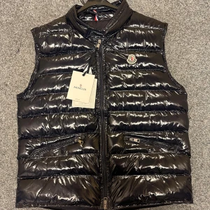 Marinblå dunväst från Moncler, blank.  - Snygg dunväst från Moncler i glansigt material med klassisk quiltad design. Västen har dragkedja framtill, två fickor med dragkedja och Moncler-logga på bröstet. Finns i svart och marinblå, perfekt för lager-på-lager och streetstyle.
