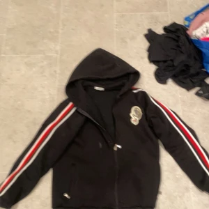 Moncler hoddie - Svart hoodiejacka med dragkedja och huva. Röda och vita ränder längs ärmarna samt ett broderat märke på bröstet. Perfekt för en sportig och avslappnad stil.