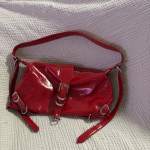 Röd axelväska med metallspännen - Cool röd axelväska i glansigt syntetmaterial med justerbar rem och flera silverfärgade metallringar och spännen. Väskan har dragkedja upptill och en dekorativ rem med spänne framtill. Perfekt för dig som gillar att sticka ut med accessoarer.