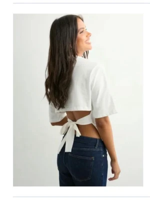 Vit croppad topp med knyt i ryggen - Säljer en vit croppad topp med korta ärmar och öppen rygg som knyts med en stor rosett baktill. Perfekt för varma dagar och ger en trendig look. Toppens material känns lätt och passar snyggt till jeans eller kjol.