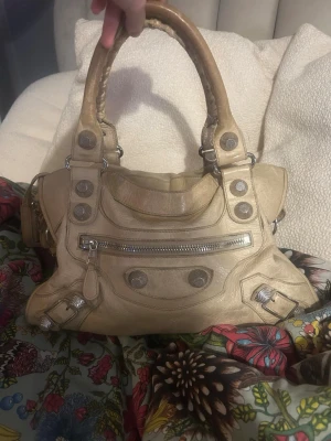 Beige Balenciaga handväska i skinn - Säljer en beige Balenciaga handväska i skinn med coola silverdetaljer och dragkedjor. Väskan rymmer ett A4! Skicket är mycket bra, dock förekommer mindre fläckar på baksidan (se bilder) och mindre slitage i några av hörnen. Självklart äkta!