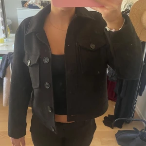 Svart kort kappa - Snygg svart kappa med stora knappar framtill och på bröstfickorna. Jackan har en boxig, croppad passform och är tillverkad i ett tjockare material som känns mjukt och värmande. Perfekt att slänga över en hoodie eller t-shirt för en chill look. Den är lite nopprig, men inget man tänker på. Priset går att diskutera!🤗