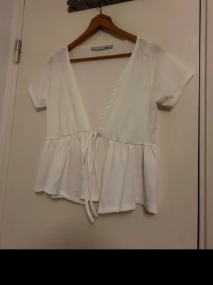 Vit omlottblus med knyt från 157 - Säljer en vit omlottblus från 157 i storlek S. Blusen har korta ärmar, v-ringad hals och knyt framtill. Den är tillverkad i ett lätt ribbat bomullstyg och har volang nedtill som ger en söt och avslappnad look. Perfekt för vår och sommar!