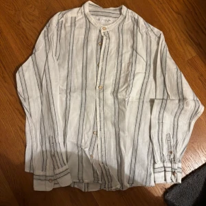 Vit randig skjorta från Zara - Vit skjorta från Zara med svarta vertikala ränder och träknappar. Skjortan har en enkel bröstficka, rund hals och långa ärmar med knapp vid manschetten. Tillverkad i ett lätt och luftigt linnematerial, perfekt för en chill och stilren look.