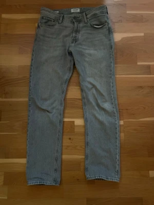 Grå jeansbyxor Jack & Jones, 30/32 - Säljer ett par klassiska grå Jack & Jones jeans i storlek 30/32. De har rak passform, fem fickor och knappgylf. Jeansen är tillverkade i slitstark denim och har den ikoniska Jack & Jones-designen på bakfickorna. Perfekt för dig som gillar tidlös stil.