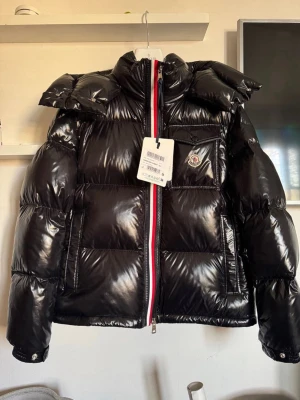 Svart pufferjacka från Moncler - Svart glansig pufferjacka från Moncler med ikonisk logga på bröstfickan och röd-vit rand vid dragkedjan. Jackan har hög krage, stora fickor och quiltad design. Materialet är polyester och dun, perfekt för kalla dagar. Snygg och modern look med långärmad passform.