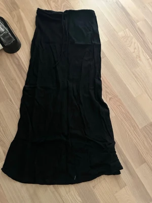 Svart långkjol med snörning - Säljer en svart långkjol med snörning i midjan. Kjolen har en enkel och stilren design, perfekt för dig som gillar en klassisk look. Materialet känns mjukt och bekvämt mot huden. Passar dig som vill ha en tidlös och mångsidig kjol.