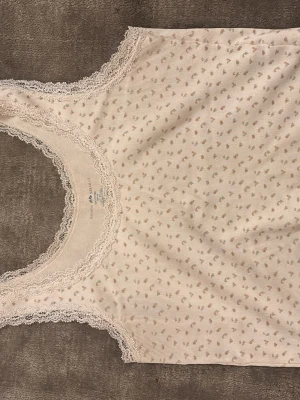 Blommig spetskantad linnetopp Brandy Melville - Supersöt ljusbeige linnetopp från Brandy Melville med små blommor i rosa och grönt. Toppen har spetskant runt halsringning och ärmhål, vilket ger en romantisk vibe. Tillverkad i 100% bomull och ärmlös, perfekt för varma dagar.