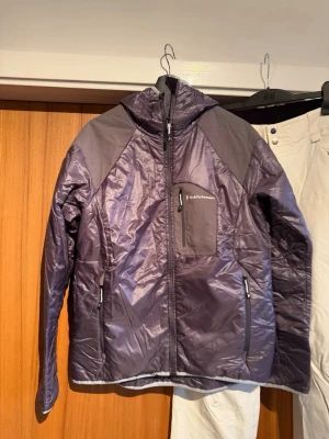 Peak performance  Primaloft jacka  - Snygg mörklila prinaloft jacka  från Peak Performance med huva och dragkedja framtill. Jackan har en praktisk bröstficka med dragkedja och två sidofickor. Materialet är glansigt och känns lätt, perfekt för kyliga dagar. Passar dig som gillar sportig och clean stil.
