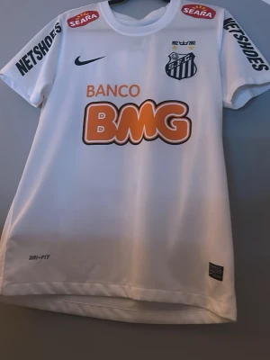 Vit Santos FC Neymar Jr matchtröja - Säljer en vit Santos FC matchtröja från Nike med Neymar Jr och nummer 11 på ryggen. Tröjan har Dri-Fit material, tryckta sponsorer och klubbmärke fram, samt coola svarta detaljer och orange BMG-logga. Perfekt för fotbollsfans som vill sticka ut på läktaren.