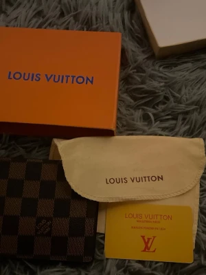 🔥NYÅRS PLÅNBOK🔥Louis Vuitton rutig plånbok - Säljer en snygg Louis Vuitton plånbok i klassiskt rutigt mönster. Plånboken är brun och svart, tillverkad i skinn med Louis Vuitton-logga. Kommer med original ask, dustbag och äkthetskort. Perfekt för dig som gillar lyxiga accessoarer.
