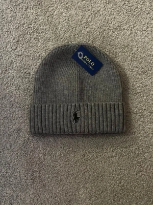 Grå stickad mössa från Polo Ralph Lauren - Säljer en snygg grå stickad mössa från Polo Ralph Lauren med klassisk ribbad kant och broderad logga framtill. Mössan har en enkel och stilren design som passar perfekt till kalla dagar. Taggen sitter kvar.