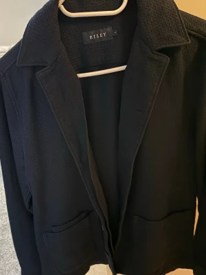 Mörkblå Riley kavaj - Säljer en mörkblå kavaj från Riley i storlek S med snygg krage och två praktiska fickor framtill. Materialet är stickat och har en lite grov struktur som ger en cool känsla. Perfekt att slänga på sig över en t-shirt eller linne när det är lite kyligt ute.