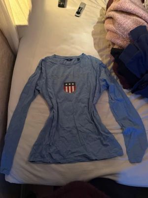 Blå långärmad topp med emblem - Säljer en blå långärmad topp med ett garn märke på bröstet Toppen har rund hals och är figursydd med normal passform. Materialet känns som mjuk bomull, perfekt för dig som gillar enkel och sportig stil.