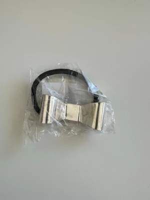 Silverfärgad hårsnodd med rosett - Snygg hårsnodd med en stor rosett i silverfärgad metall och svart elastiskt band. Perfekt för att ge frisyren en cool och stilren touch. Rosetten har en blank finish och är riktigt statement. Passar dig som gillar att sticka ut med accessoarer.