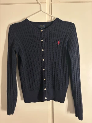 Marinblå kofta från Polo Ralph Lauren - Snygg marinblå kofta från Polo Ralph Lauren, sitter som storlek S/M. Den är kabelstickad med vita knappar framtill och har det klassiska röda Polo-logot broderat på bröstet. 