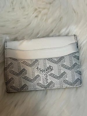 Vit plånbok - Säljer en stilren vit G plånbok med den ikoniska grafiska mönster i svart och grått. Plånboken har flera kortfack och är tillverkad i skinn. Perfekt för dig som gillar exklusiva accessoarer med unik design.