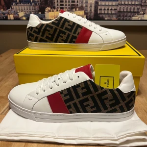 Fendi vita sneakers med röd detalj - Snygga vita sneakers från Fendi med röd läderdetalj på plösen och sidan. Ena sidan har Fendis klassiska logomönster i svart och brunt. Skorna har rund tå, platt sula och snörning. Tillverkade i skinn och textil för en lyxig känsla.