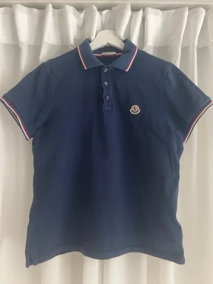 Marinblå Moncler pikétröja (äkta) - Snygg marinblå pikétröja från Moncler med klassisk krage och korta ärmar. Tröjan har vita och röda ränder på krage och ärmslut samt Moncler-logga på bröstet. Köpt på NK, nypris 3600. Skick 8/10 inga synliga defekter som till exempel hål. Storlek S, säljer den då den har blivit för liten. Kontakta mig om du har några frågor!