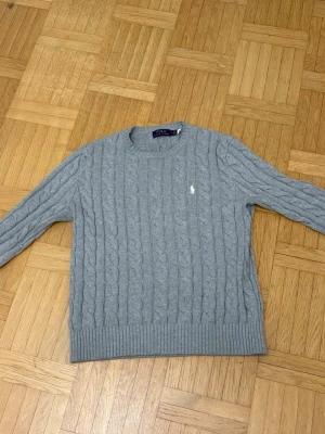 Grå kabelstickad tröja Polo Ralph Lauren - Snygg grå kabelstickad tröja från Polo Ralph Lauren med klassisk rund hals och långärmad passform. Tröjan har det ikoniska vita broderade logot på bröstet och ribbade muddar vid ärmslut och nederkant. Perfekt för dig som gillar stilrena och tidlösa plagg.