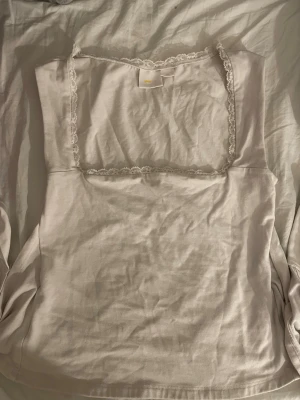 Vit omlottopp med spetskant - Säljer en vit omlottopp i bomull med fyrkantig ringning och dekorativ spetskant runt halsen. Toppen är ärmlös och har öppen rygg med knytband nedtill för en snygg detalj. Perfekt för dig som gillar stilrena och feminina plagg.