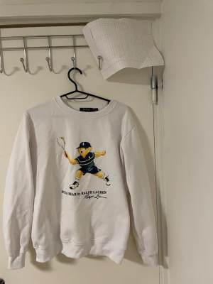 Vit sweatshirt Polo Bear Ralph Lauren - Vit sweatshirt från Polo Ralph Lauren med ikoniskt Polo Bear-tryck där björnen spelar tennis. Tröjan har rund hals, långa ärmar och ribbade muddar. Perfekt för dig som gillar sportiga och klassiska streetwear-vibbar.Liknande modell som orginala. 