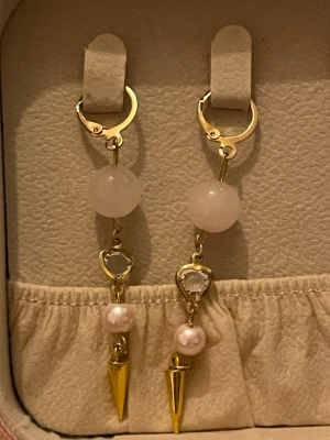 Guldiga örhängen med pärlor och spetsdetalj - Säljer ett par snygga örhängen i guldton med rund vit pärla, hjärtformad transparent detalj och spetsig guldpendel längst ner. Perfekt för dig som gillar att sticka ut med accessoarer! Lätta att sätta i med smidig låsning.  6cm lång