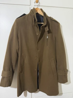 Brun rock från Mustal by Daas, M - Snygg brun rock från Mustal by Daas i storlek M. Jackan har klassisk krage, axelklaffar, knappstängning och en bröstficka med dragkedja. Fodrad insida och detaljer vid ärmslut. Perfekt för dig som gillar stilrena och tidlösa plagg.