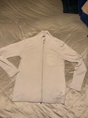 Beige kofta med dragkedja j.Lindberg  - Säljer en stilren beige kofta från j.Lindbergmed hel dragkedja framtill och en liten bröstficka. Koftan har långärmade ärmar och är gjord i 100%  merinoull. Enkel och clean design som passar till det mesta.