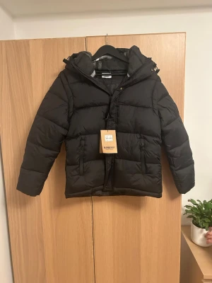 Svart pufferjacka från Burberry - Säljer en svart pufferjacka från Burberry med huva och dragkedja. Jackan har ett stort Burberry London England-märke på ärmen och rutigt foder i huvan. Perfekt för kalla dagar och riktigt snygg till streetwear-stilen. Kan även användas som väst.