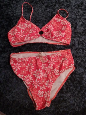 Blommig bikini från SHEIN - Säljer en söt bikini från SHEIN i storlek S. Bikinin är rosa med vita blommor och har ett ringformat hål mellan kuporna. Axelbanden är smala och trosan är högt skuren. Perfekt för strandhäng och poolparty!